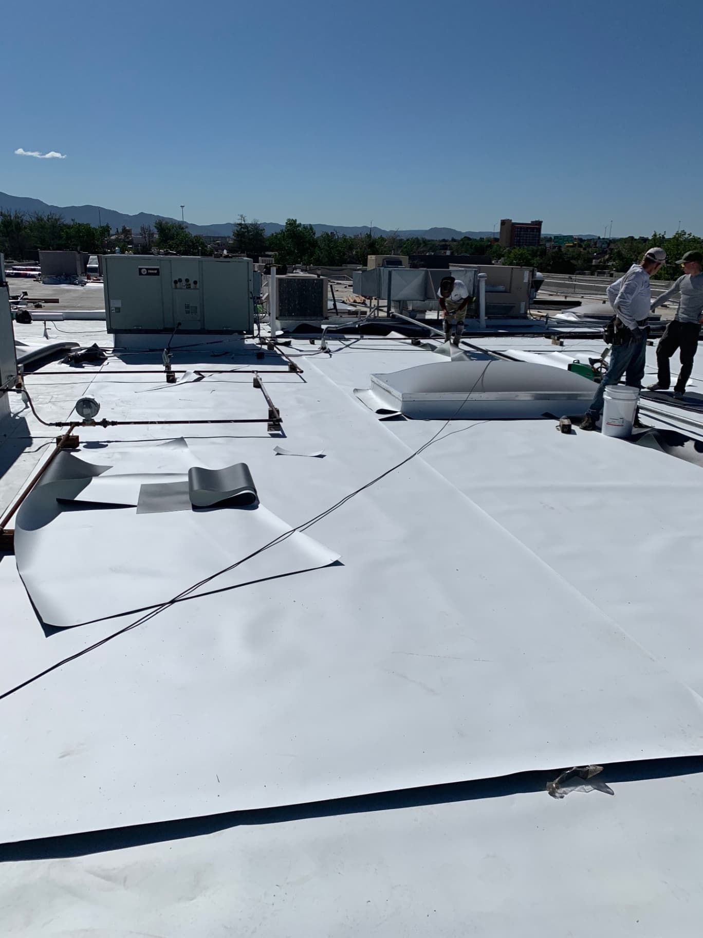 Roofing crew laying out and fastening white TPO membrane around skylights and rooftop units